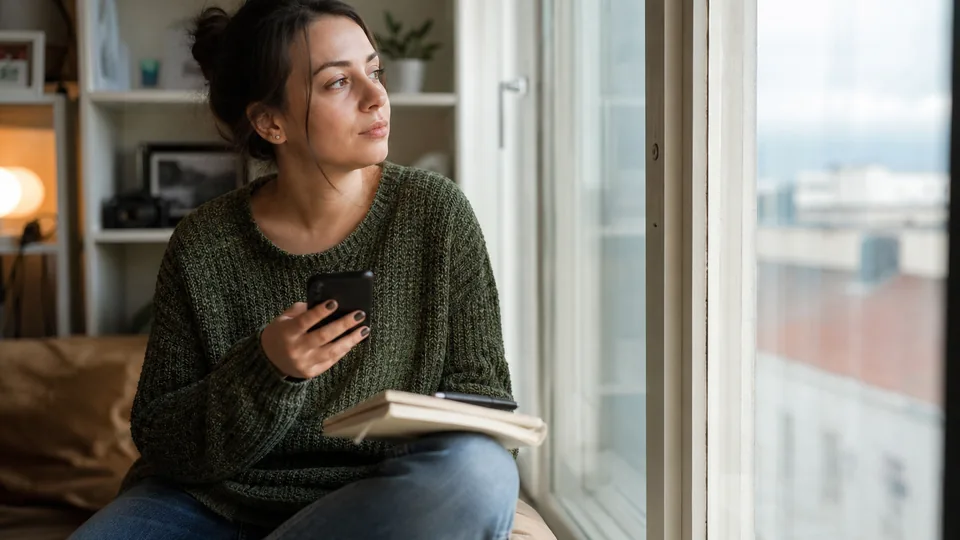 Adult woman on a window seat holding her phone and weighing two companion app subscriptions