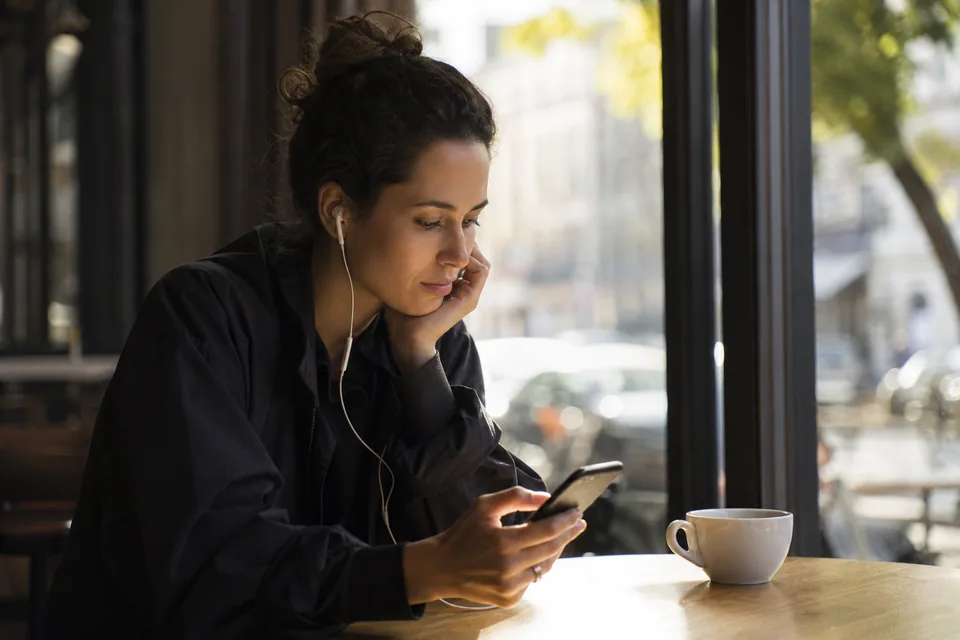 Adult woman at a cafe window with earbuds, checking a chat app subscription on her phone