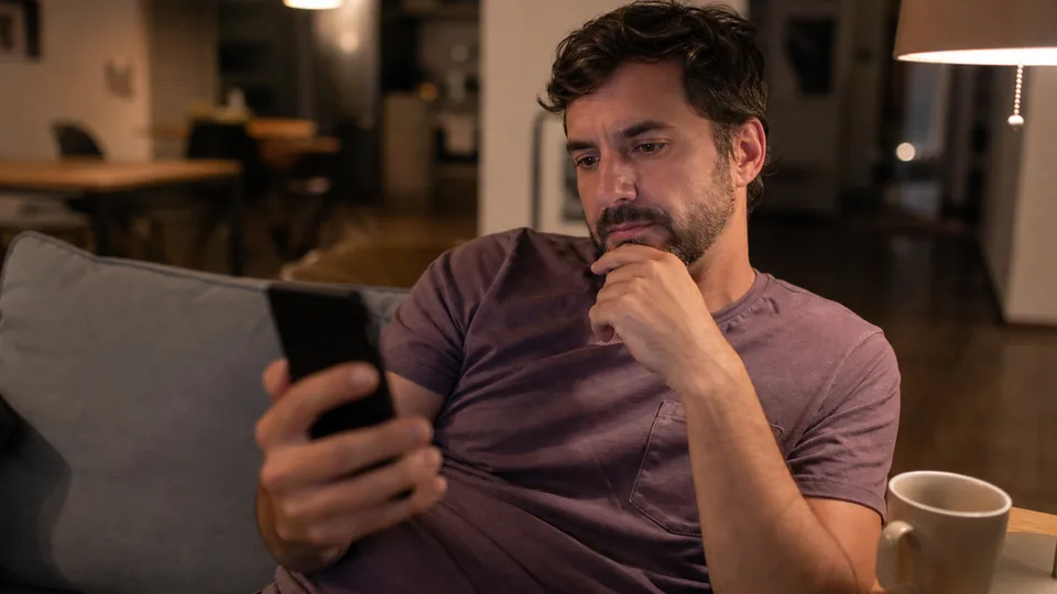 Adult man on a sofa in the evening reading an app subscription price list on his phone